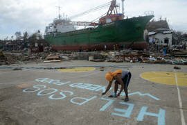 Seorang pria mengecat pesan bertuliskan "Tolong SOS Kami Perlu Makanan” di lapangan basket di depan sebuah kapal yang terdampar di Anibong, Tacloban, Senin (11 November). Tiga hari sebelumnya, Topan Super Haiyan menghantam negara itu, menewaskan sekitar 10.000 orang, menyebabkan jutaan orang kehilangan rumah dan menyebabkan kerugian senilai ratusan juta dolar. Amerika Serikat dan PBB memobilisasi bantuan darurat sementara besarnya kehancuran ini mulai terlihat. [Noel Celis/AFP]